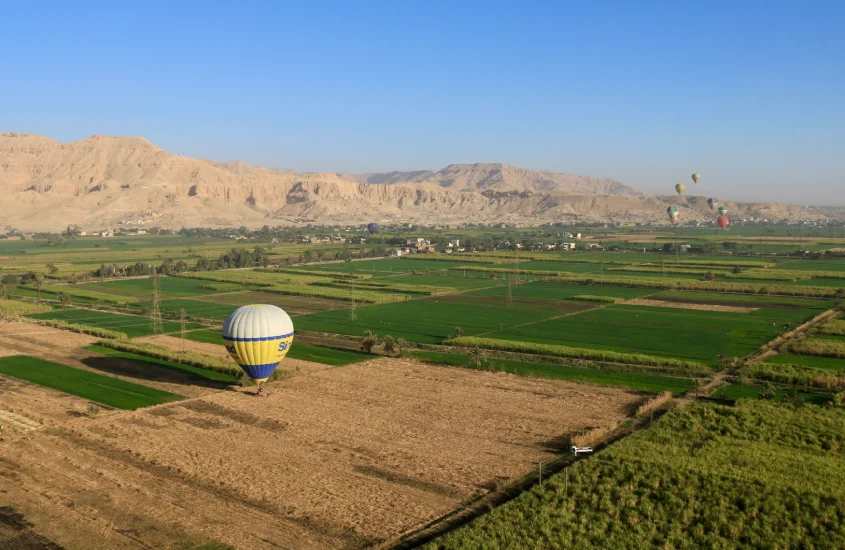 Vista aérea de um balão de ar quente pairando sobre uma paisagem agrícola, com campos verdes e montanhas ao fundo sob um céu azul claro.