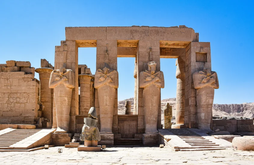 Estrutura antiga com colunas e estátuas monumentais em Luxor, esculpidas em pedra, representando a grandiosidade da arquitetura egípcia antiga sob um céu azul brilhante.