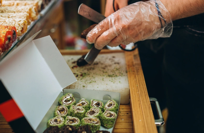 Mão de uma pessoa preparando doces locais, usando pinças para cortar e organizar os doces em uma caixa, capturando a tradição e a arte da culinária.