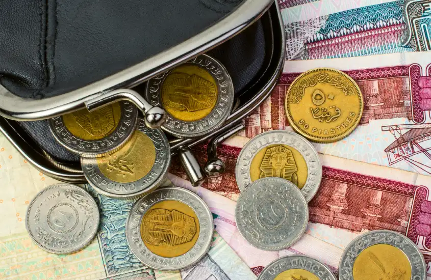 Moedas egípcias e notas espalhadas em torno de uma pequena bolsa de couro, mostrando a moeda local com detalhes de figuras históricas.