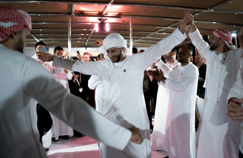 Homens vestindo trajes tradicionais árabes dançam alegremente em um círculo, celebrando em uma festa cultural com sorrisos e entusiasmo.