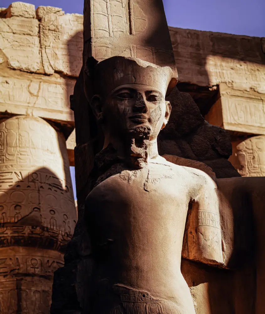 Grande estátua esculpida de um faraó egípcio em um templo ao ar livre, iluminada pela luz suave do fim da tarde, destacando a imponência dos detalhes históricos.