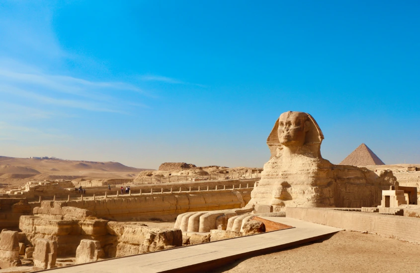 A Esfinge majestosa no deserto de Gizé, com o céu azul brilhante ao fundo e uma das pirâmides visíveis à distância.