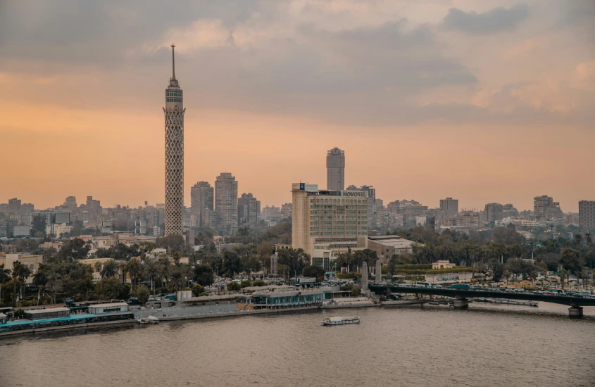 Vista do Cairo ao entardecer, com a famosa Torre do Cairo se destacando no horizonte e o rio Nilo em primeiro plano.