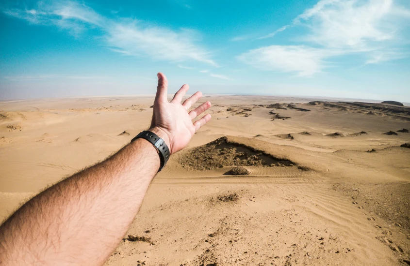 Uma mão estendida em direção à vasta extensão de um deserto, sob um céu azul com algumas nuvens.