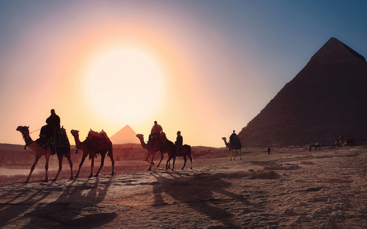 Pessoas montadas em camelos atravessando o deserto ao entardecer, com as pirâmides ao fundo e o sol iluminando tudo em tons quentes.