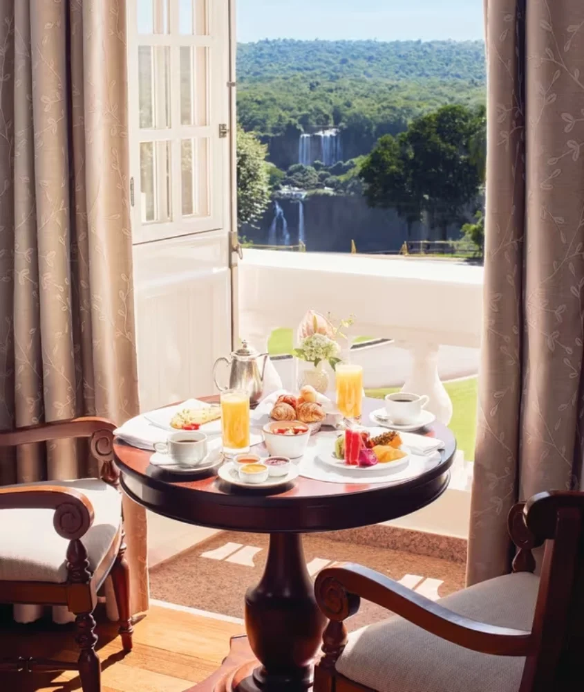 Café da manhã servido em uma mesa junto à janela aberta, com vista direta para as impressionantes Cataratas do Iguaçu, cercadas pela floresta.