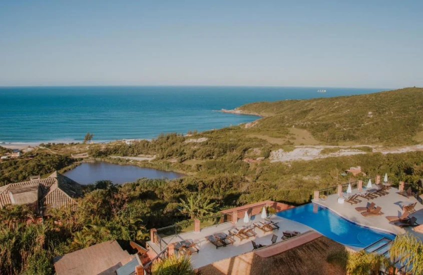 Vista panorâmica de uma piscina infinita e uma extensa faixa de praia, cercada por uma vegetação exuberante, com o mar ao fundo.