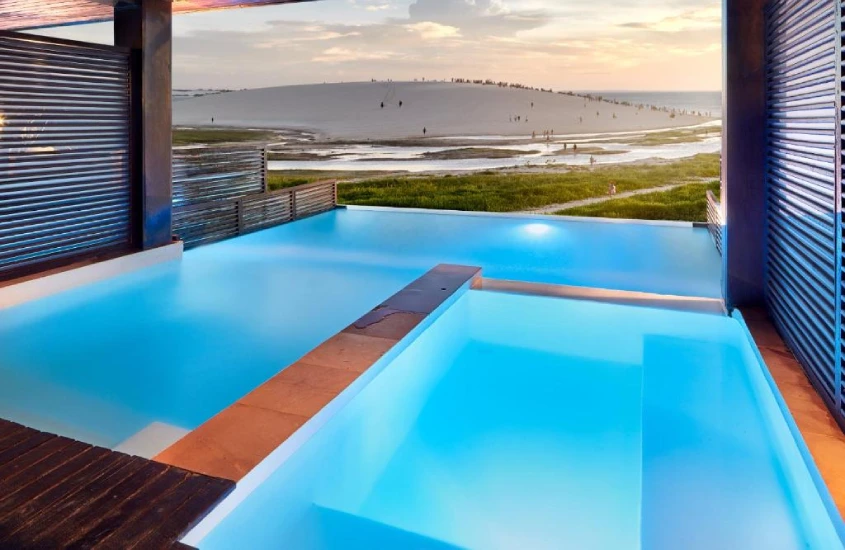 Piscina infinita com borda de vidro, oferecendo uma vista deslumbrante para as dunas ao pôr do sol, com um céu alaranjado ao fundo.