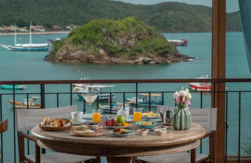 Mesa ao ar livre com café da manhã servido, vista para a baía e barcos ancorados em uma área tranquila, cercada por montanhas verdes ao fundo.