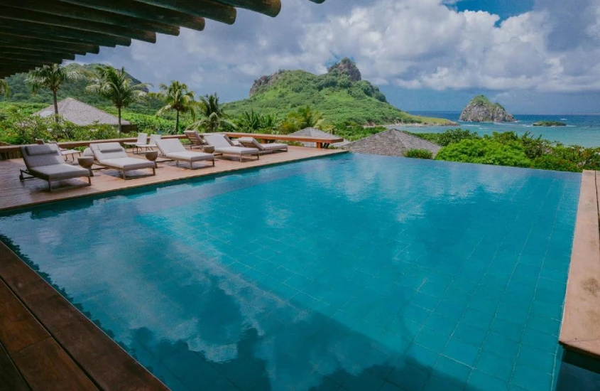 Uma piscina de borda infinita com espreguiçadeiras ao redor, cercada por vegetação tropical e com vista para formações rochosas e o mar azul ao fundo.