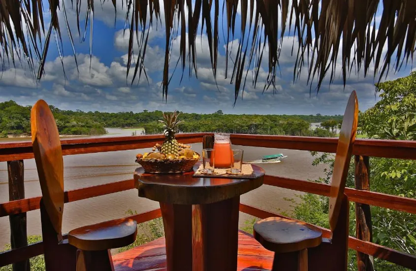 Varanda charmosa com uma mesa rústica de madeira e duas cadeiras, cercada por vegetação tropical, com vista para um rio na Amazônia e um barco ao fundo.