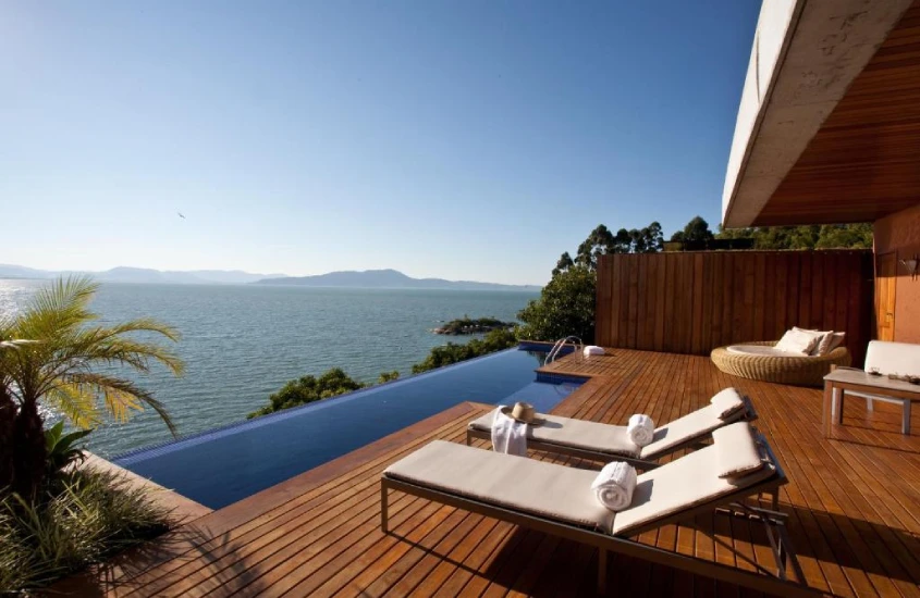 Duas espreguiçadeiras de madeira ao lado de uma piscina de borda infinita, com vista para o mar tranquilo e céu claro, cercado por vegetação costeira.