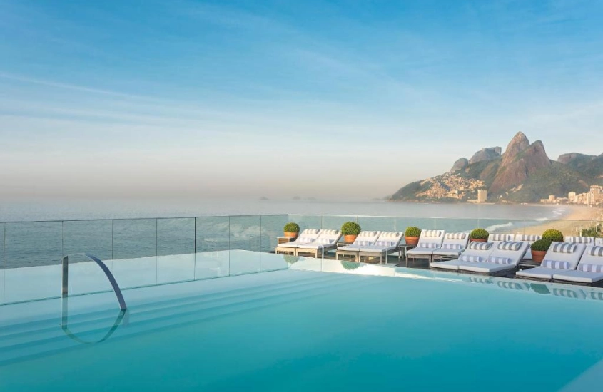 Piscina na cobertura com vista deslumbrante para a praia de Ipanema e o Morro Dois Irmãos ao fundo, em um cenário de céu azul claro.