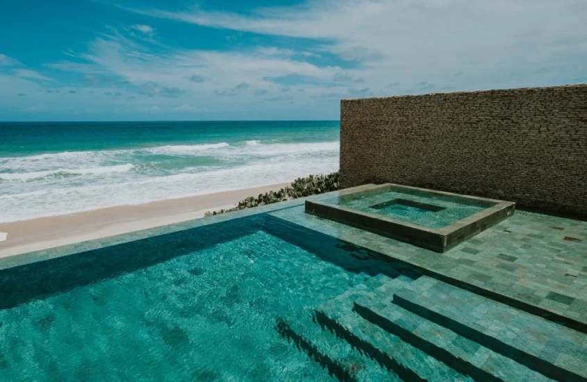 Piscina de borda infinita com jacuzzi, de frente para o mar, cercada por pedras e uma parede de tijolos rústicos.