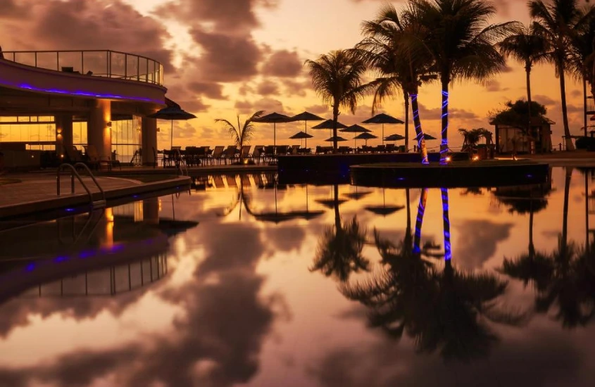 Reflexo das palmeiras e do céu alaranjado de um pôr do sol em uma piscina ao ar livre iluminada por luzes azuis, com espreguiçadeiras ao redor.