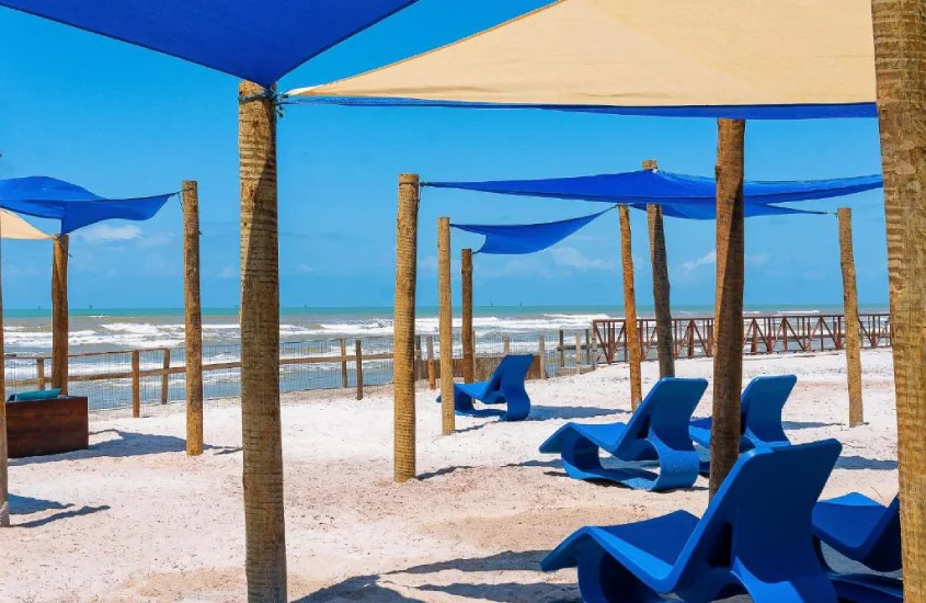 Espreguiçadeiras azuis em um espaço de praia coberto por tendas de tecido, com vista para o mar em um dia de céu limpo.