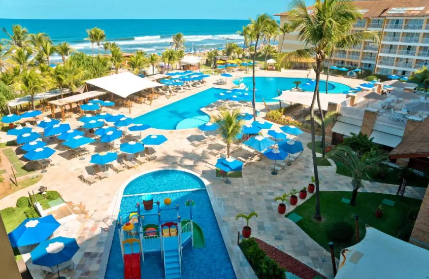 Uma vista aérea de piscinas ao ar livre, rodeadas por guarda-sóis azuis e palmeiras, com o mar ao fundo.