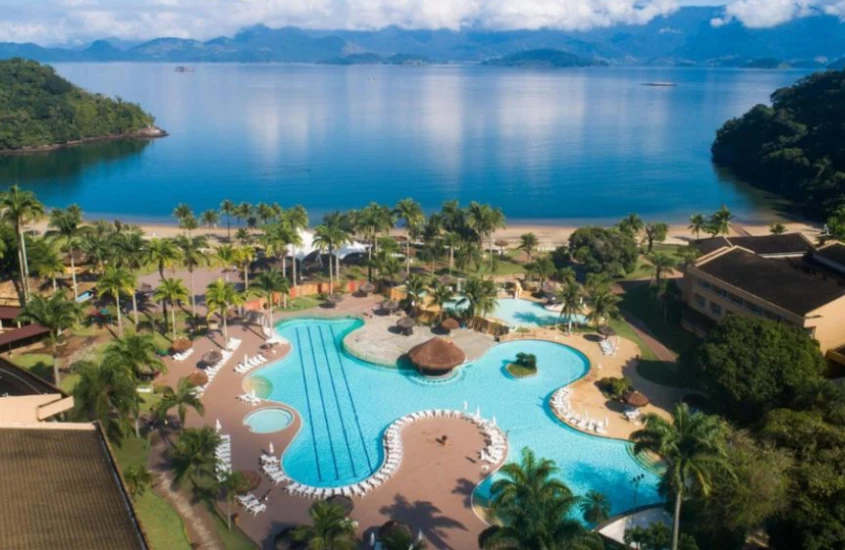 Vista panorâmica de um resort com piscina e praia em um cenário de montanhas e mar, capturada em plena luz do dia