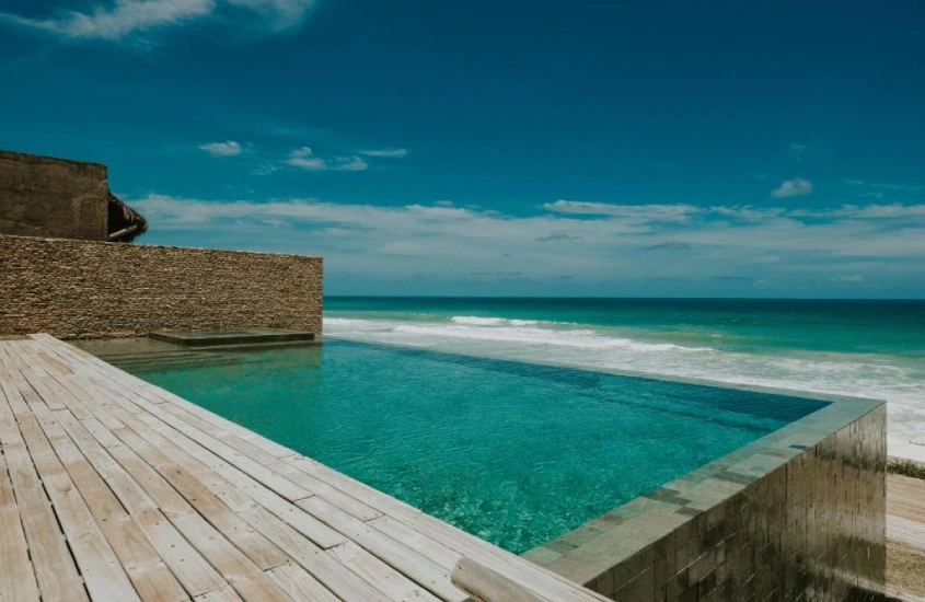 Uma piscina de borda infinita de frente para o mar, cercada por madeira rústica e com o céu azul claro ao fundo.