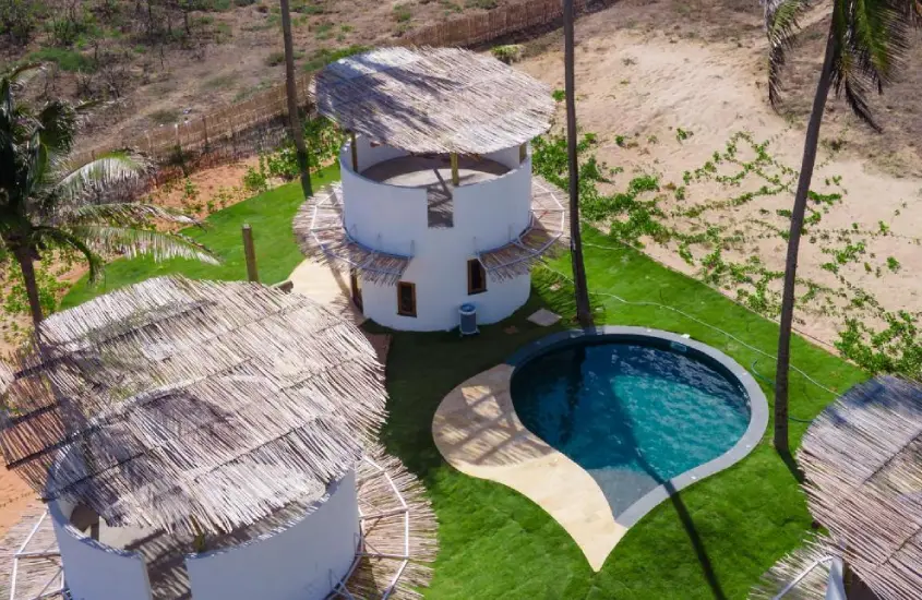 Vista aérea de estruturas circulares de palha ao lado de uma piscina em formato orgânico, rodeada por gramado e coqueiros em um ambiente rústico à beira da praia.