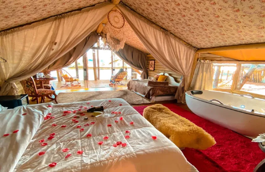 Interior de uma luxuosa tenda de glamping, com cama decorada com pétalas de rosa e uma banheira de hidromassagem em destaque.