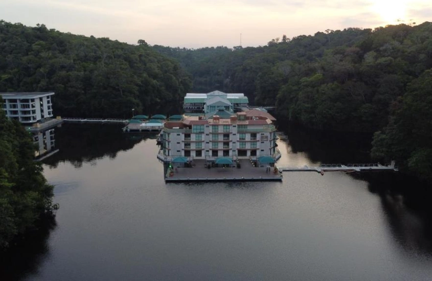 Um grande hotel flutuante ao entardecer, cercado pela floresta amazônica, em uma paisagem serena refletida nas águas escuras ao redor.