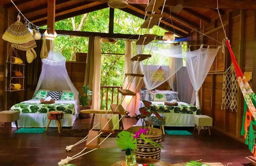 Um quarto rústico, porém aconchegante, decorado com tons naturais e redes, em uma casa de madeira imersa na floresta tropical, cercada por árvores.