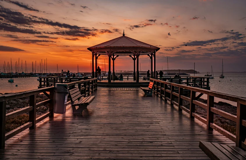 Vista de um pôr do sol no pier de madeira com um quiosque ao fundo e barcos ancorados no horizonte