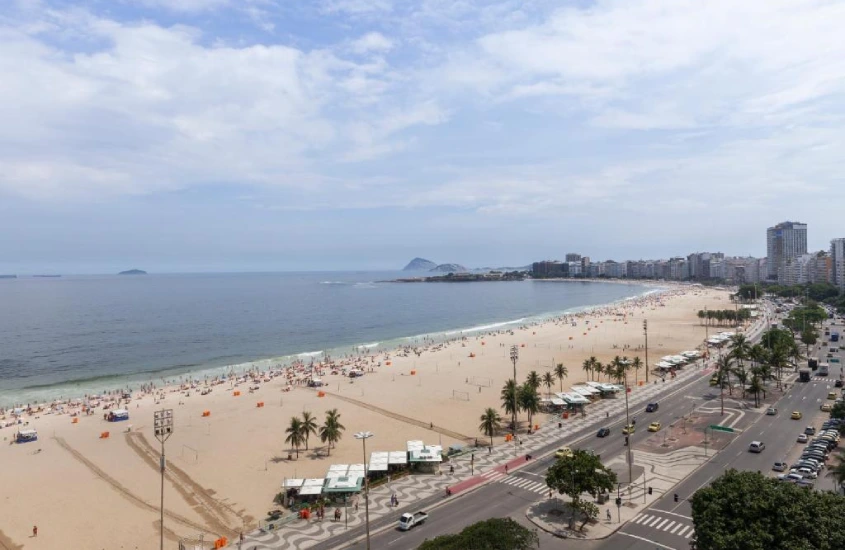 Vista aérea da Praia de Copacabana, com a faixa de areia cheia de guarda-sóis, o calçadão movimentado e o mar azul ao fundo.