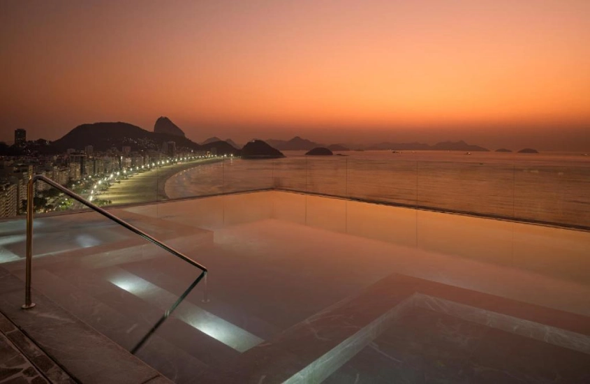 Piscina de borda infinita iluminada ao entardecer, com uma vista espetacular para a Praia de Copacabana e o Pão de Açúcar ao fundo.