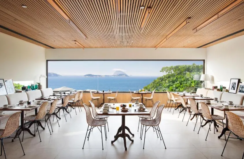 Restaurante com mesas de madeira e cadeiras de vime, com uma ampla janela oferecendo vista para o mar e o horizonte.