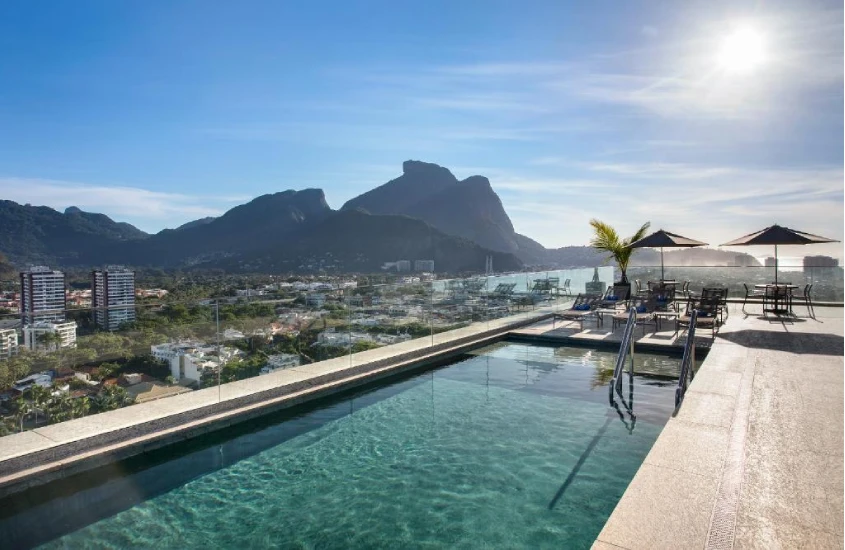 Piscina com espreguiçadeiras e guarda-sóis no rooftop, com uma vista para as montanhas e o mar da Barra da Tijuca.