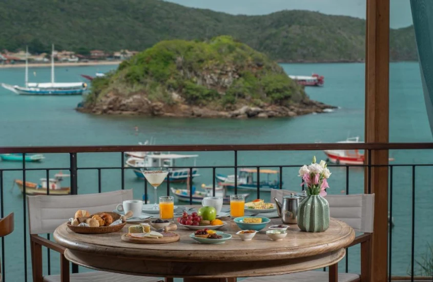 Mesa de café da manhã em uma varanda, com vista para o mar e pequenas ilhas ao fundo, em um cenário ensolarado.