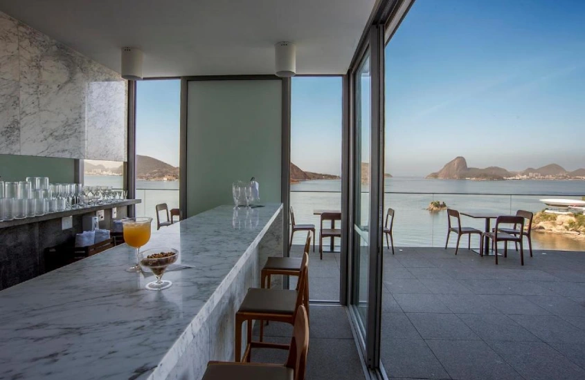 Bar com balcão de mármore e janelas amplas de vidro, oferecendo vista para o Pão de Açúcar e a Baía de Guanabara.