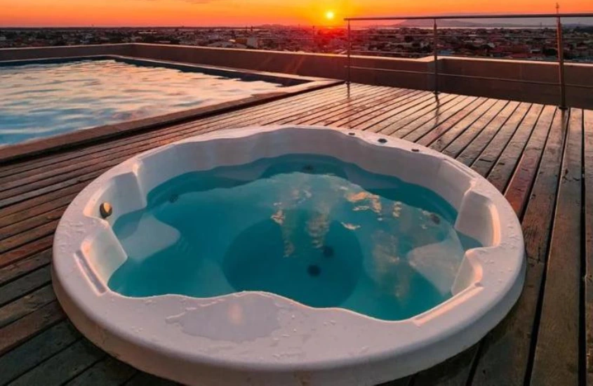 Jacuzzi e piscina no rooftop ao pôr do sol, com céu alaranjado e vista para a cidade ao fundo.