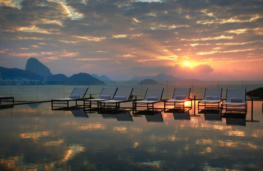 Uma piscina de borda infinita ao pôr do sol, com espreguiçadeiras de frente para o Pão de Açúcar e o mar, refletindo o céu alaranjado.