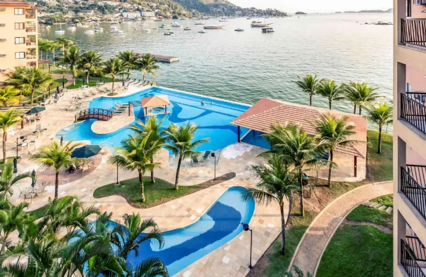 Piscinas ao ar livre rodeadas por coqueiros, à beira da Baía de Angra dos Reis, com o mar ao fundo em um cenário paradisíaco.