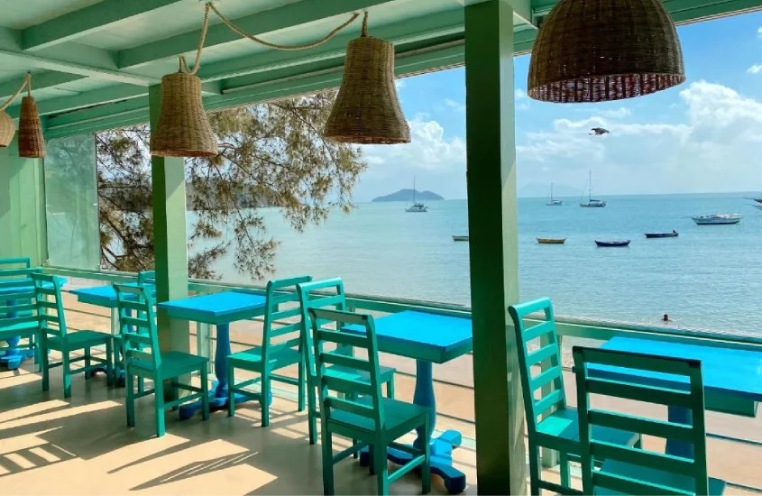 Varanda decorada em tons de azul e verde, com mesas e cadeiras voltadas para o mar, barcos e montanhas ao fundo em Búzios.