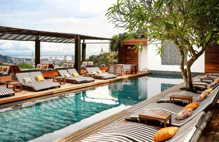 Espreguiçadeiras dispostas ao redor de uma piscina com estrutura de madeira e vegetação, com vista para o bairro de Santa Teresa e o verde ao redor.