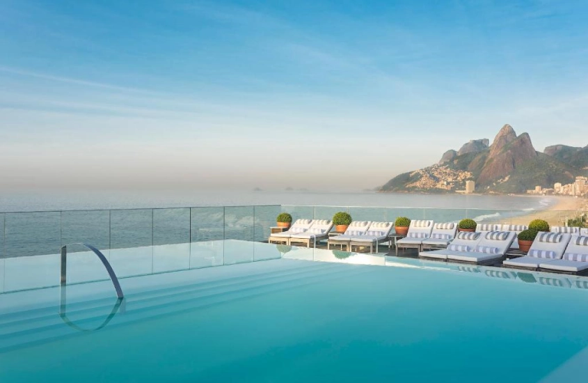 Piscina de borda infinita no rooftop com vista para o mar e o Morro Dois Irmãos, em um cenário ensolarado com espreguiçadeiras ao lado.