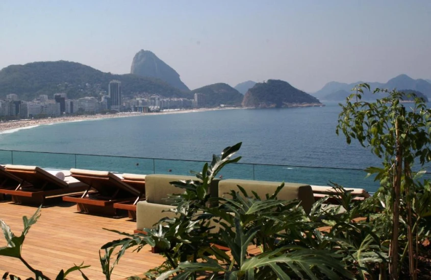 Piscina de vidro transparente no rooftop, com espreguiçadeiras ao redor e vista para o mar de Copacabana, cercada por uma paisagem urbana.