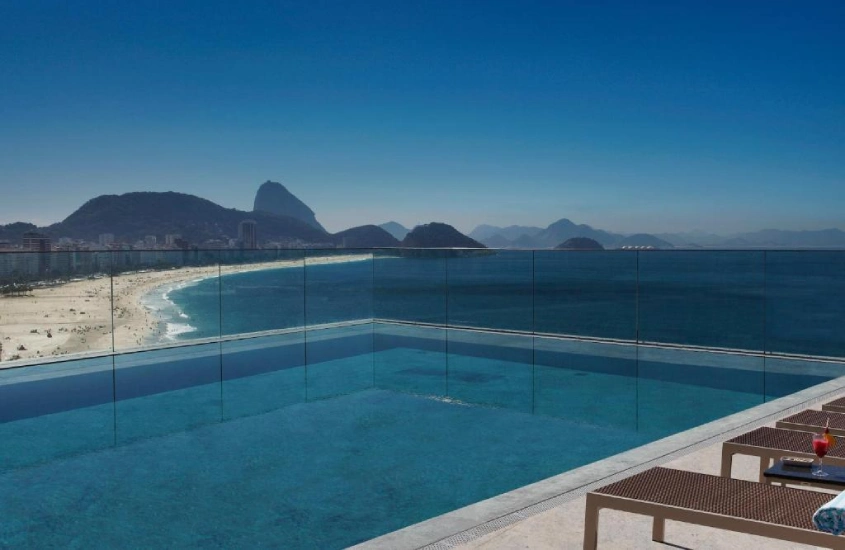 Piscina na cobertura com vista para a Praia de Copacabana, rodeada por espreguiçadeiras e com o mar ao fundo, em um dia de céu limpo.