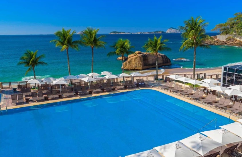 Grande piscina ao ar livre com palmeiras ao redor, de frente para o mar e cercada por cadeiras de praia, em um cenário tropical.