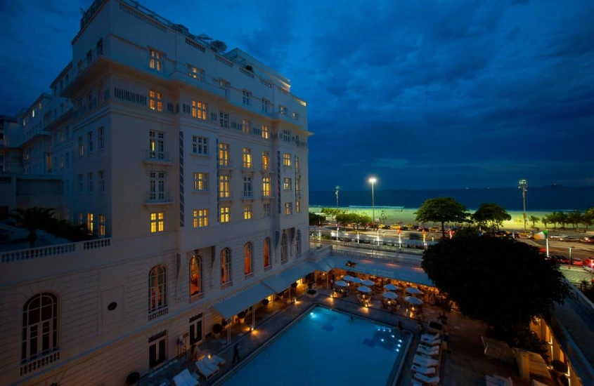 Fachada histórica do Copacabana Palace à noite, com luzes acesas, piscina iluminada e vista para a Praia de Copacabana ao fundo.