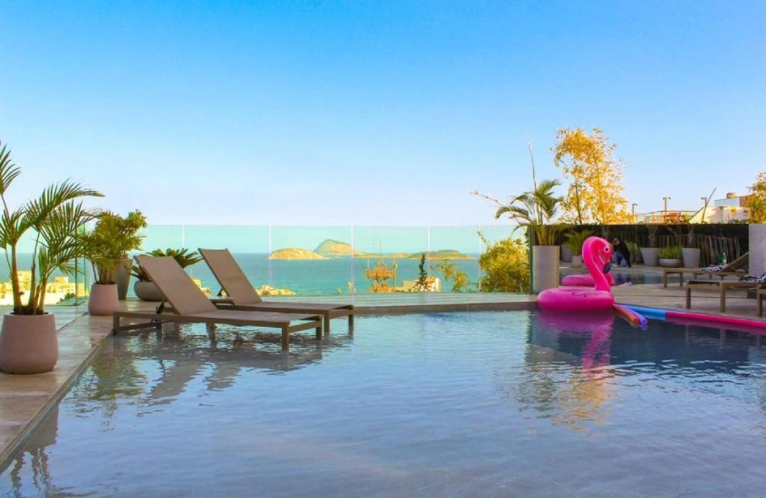 Piscina no rooftop, cercada por espreguiçadeiras e vegetação tropical, com vista para a Praia de Copacabana em um ambiente tranquilo.