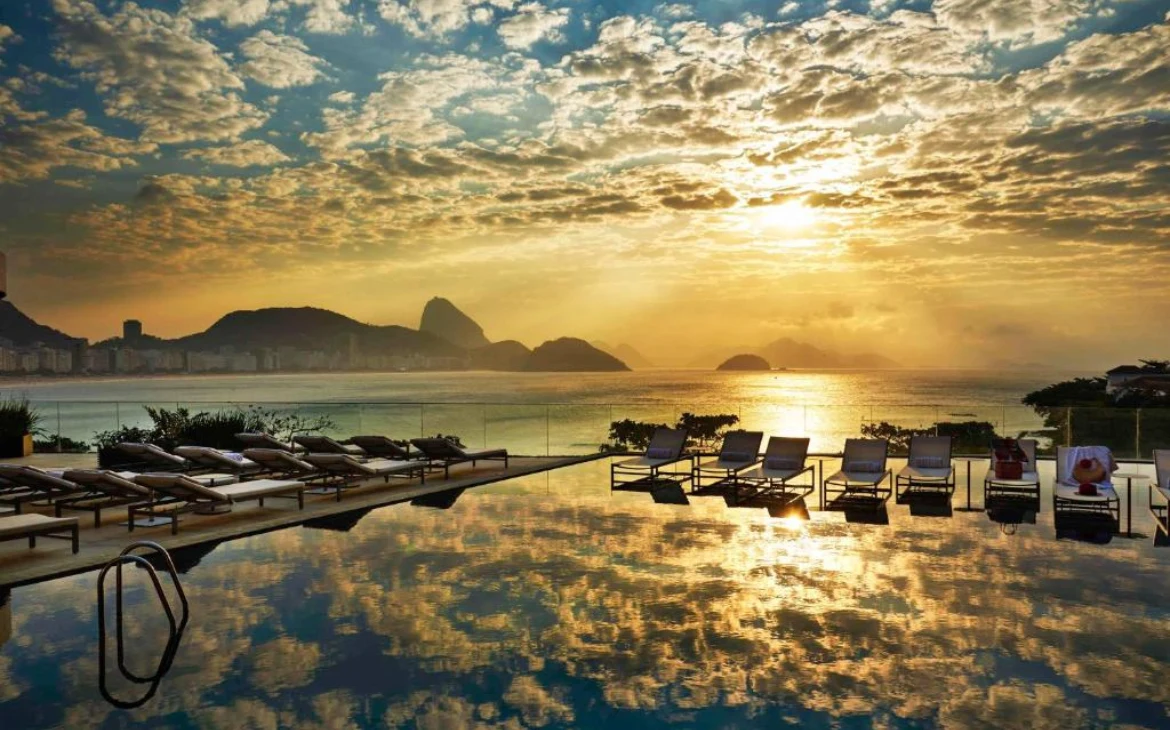 Uma piscina de borda infinita ao entardecer, com espreguiçadeiras ao redor e reflexo do céu alaranjado, oferecendo uma vista panorâmica para o Pão de Açúcar e o mar