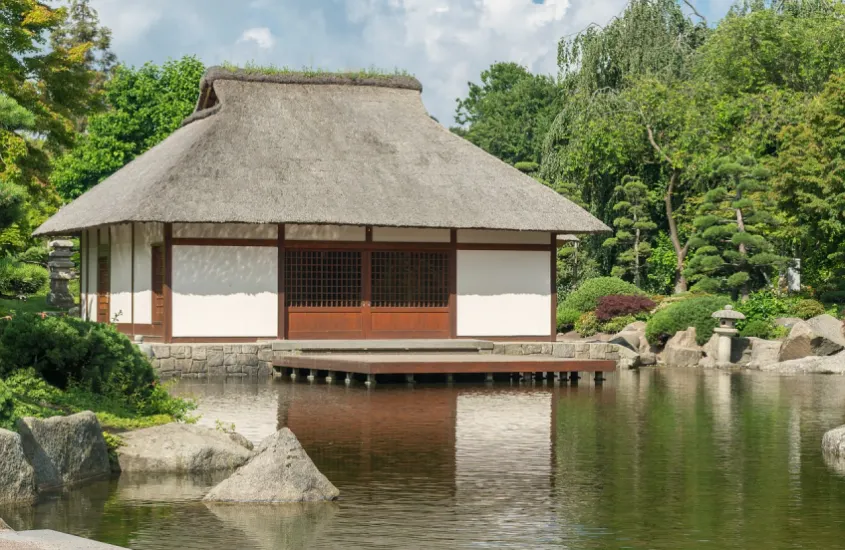 Estrutura tradicional japonesa com telhado de palha e paredes de madeira ao lado de um lago sereno e rodeada por vegetação verdejante.