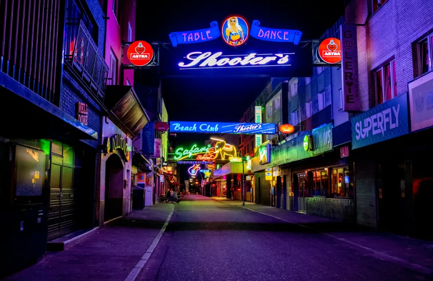 Corredor estreito e vibrante de uma rua com várias placas de neon coloridas anunciando bares e clubes noturnos.