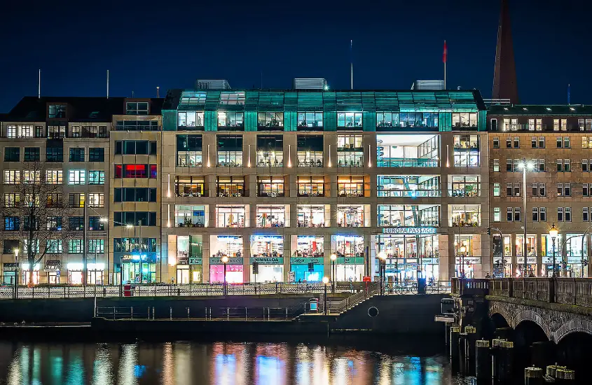 Fachada do centro comercial Europa Passage com janelas iluminadas e reflexos no canal à noite.